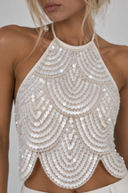 A close-up of a model wearing a halter-style crop top covered in shimmering silver sequins arranged in a scalloped, fan-like pattern against a light gray studio background.